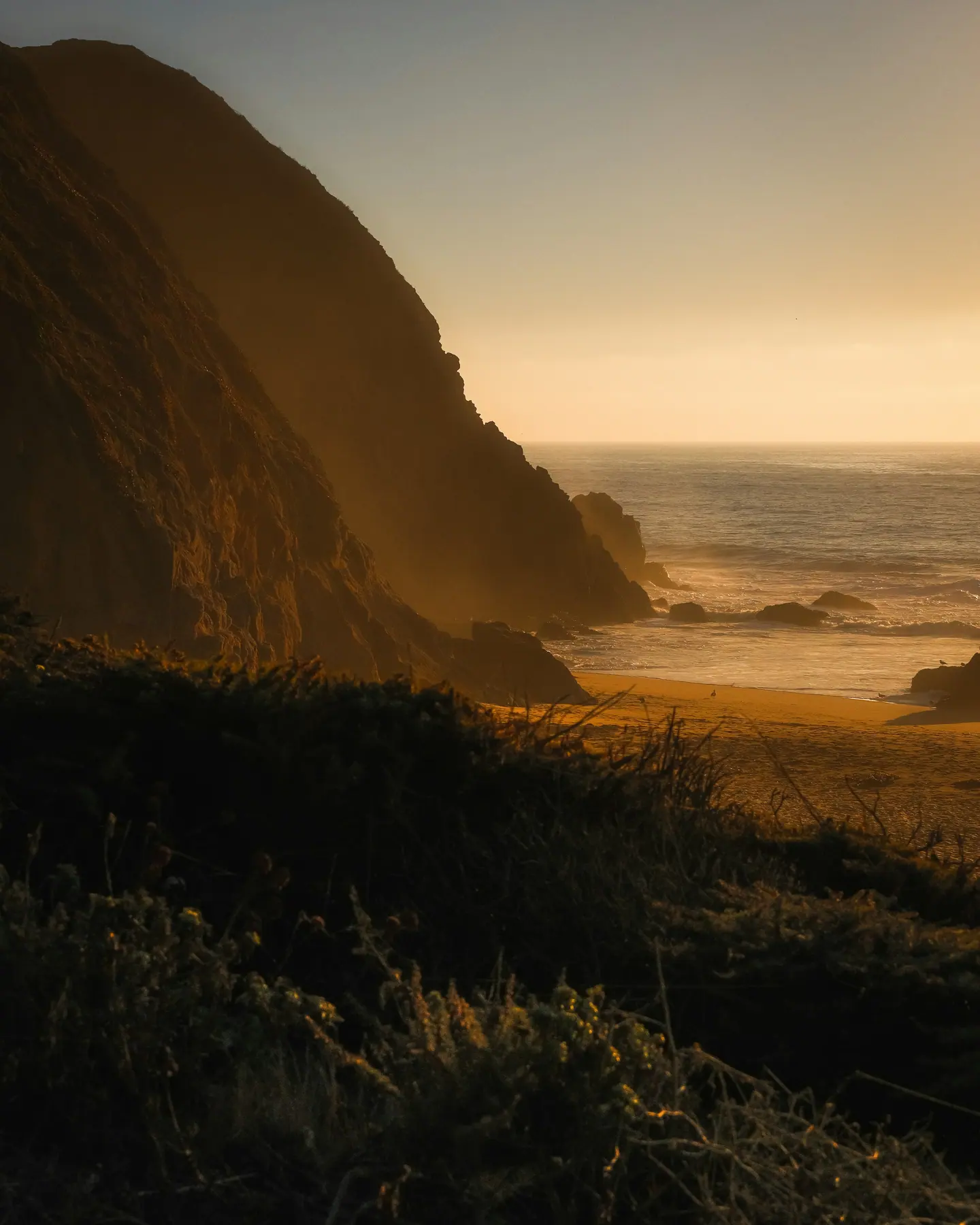 A hazy coastline at golden hour, cliffs falling to an empty beach.
